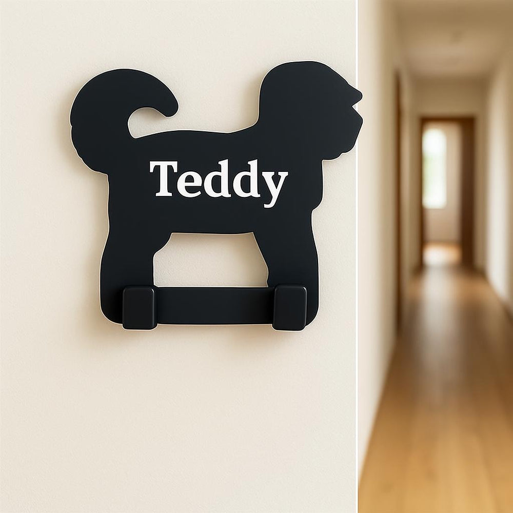 Black dog-shaped hook with 'Teddy' on a white wall, blurred hallway in the background