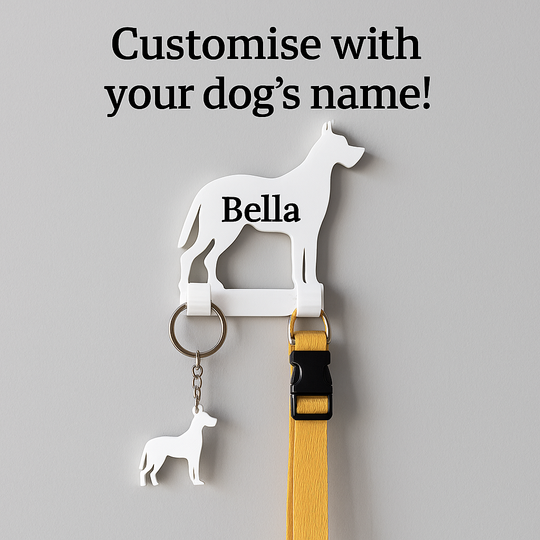 White 3D-printed Great Dane wall hook personalised with the name 'Bella', mounted on a light grey wall. The hook holds a yellow dog lead and a matching dog-shaped keyring. Bold black text above reads 'Customise with your dog’s name!' promoting personalisation options.