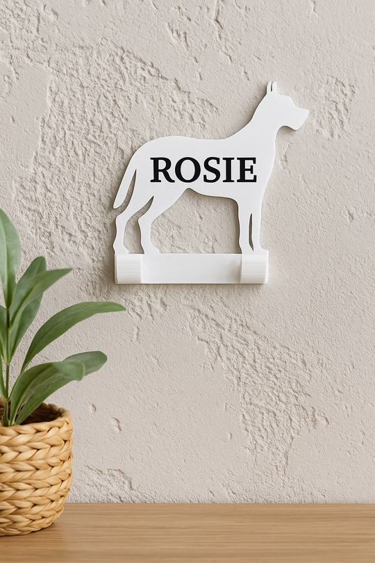 White 3D-printed Great Dane-shaped wall hook personalised with the name 'ROSIE' in bold black letters. Mounted on a textured off-white wall above a wooden surface with a woven plant pot, creating a clean, modern entryway look.