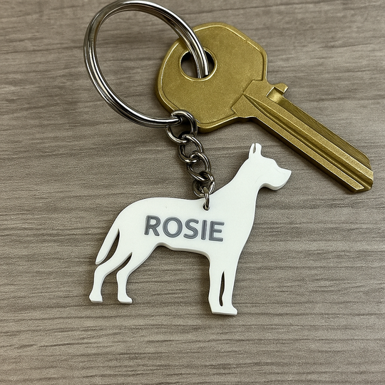 White 3D-printed Great Dane-shaped keyring personalized with the name 'Rosie'. Attached to a silver keyring holding 1 house keys, shown on a wooden background. Minimalist custom pet accessory for dog lovers.